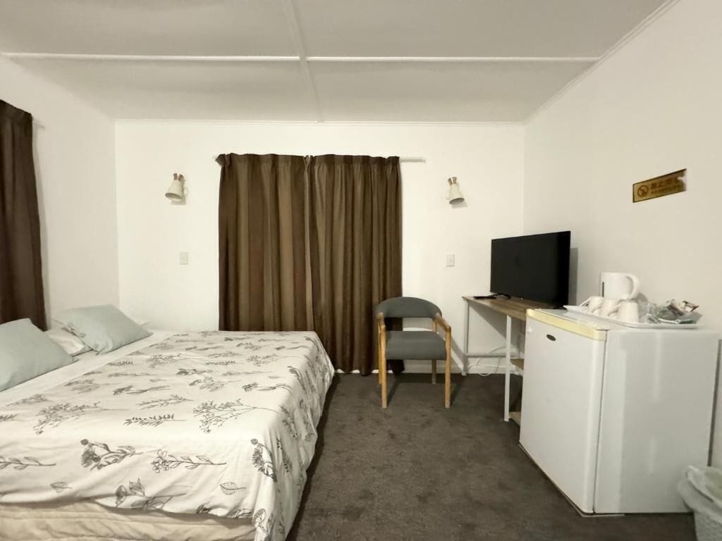 A hotel room with a bed, a desk with a television, a chair, and a small refrigerator.