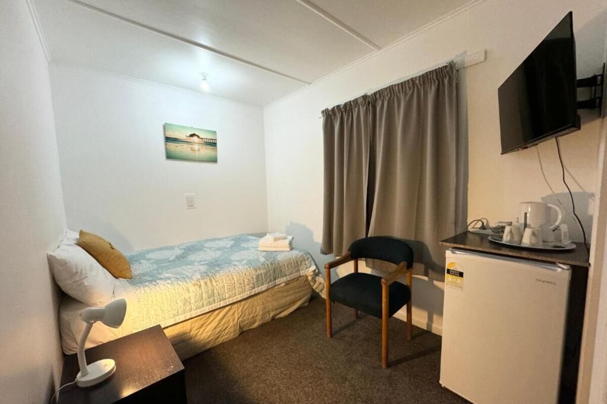 Ett litet, välordnat rum med en säng, en stol, ett skrivbord, en tv och ett kylskåp.