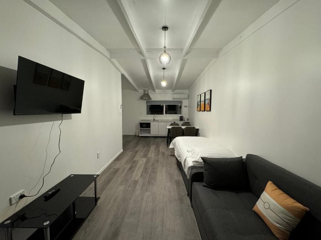 A modern hotel room with a flat-screen TV, a sofa, a desk, and a kitchenette in the background.