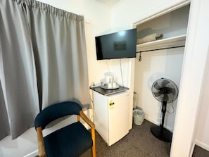A compact room with a bed, a small white cabinet, a television mounted on the wall, a fan, and a chair.