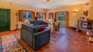 A spacious living room with a sofa, armchairs, a coffee table, and a fireplace. There are large windows and a door leading outside.