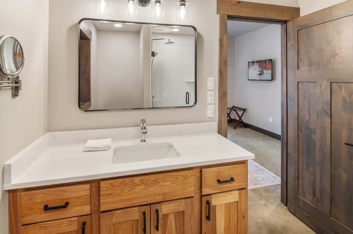Une salle de bain avec un lavabo blanc, des meubles en bois, un grand miroir et une porte en bois.