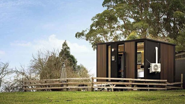 Une petite cabane en bois sombre, dotée d’un porche et d’une porte, entourée d’une clôture en bois et d’arbres.
