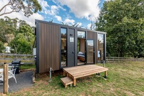 Una cabaña moderna y compacta con una terraza de madera y una pequeña mesa, rodeada de paisajes naturales.