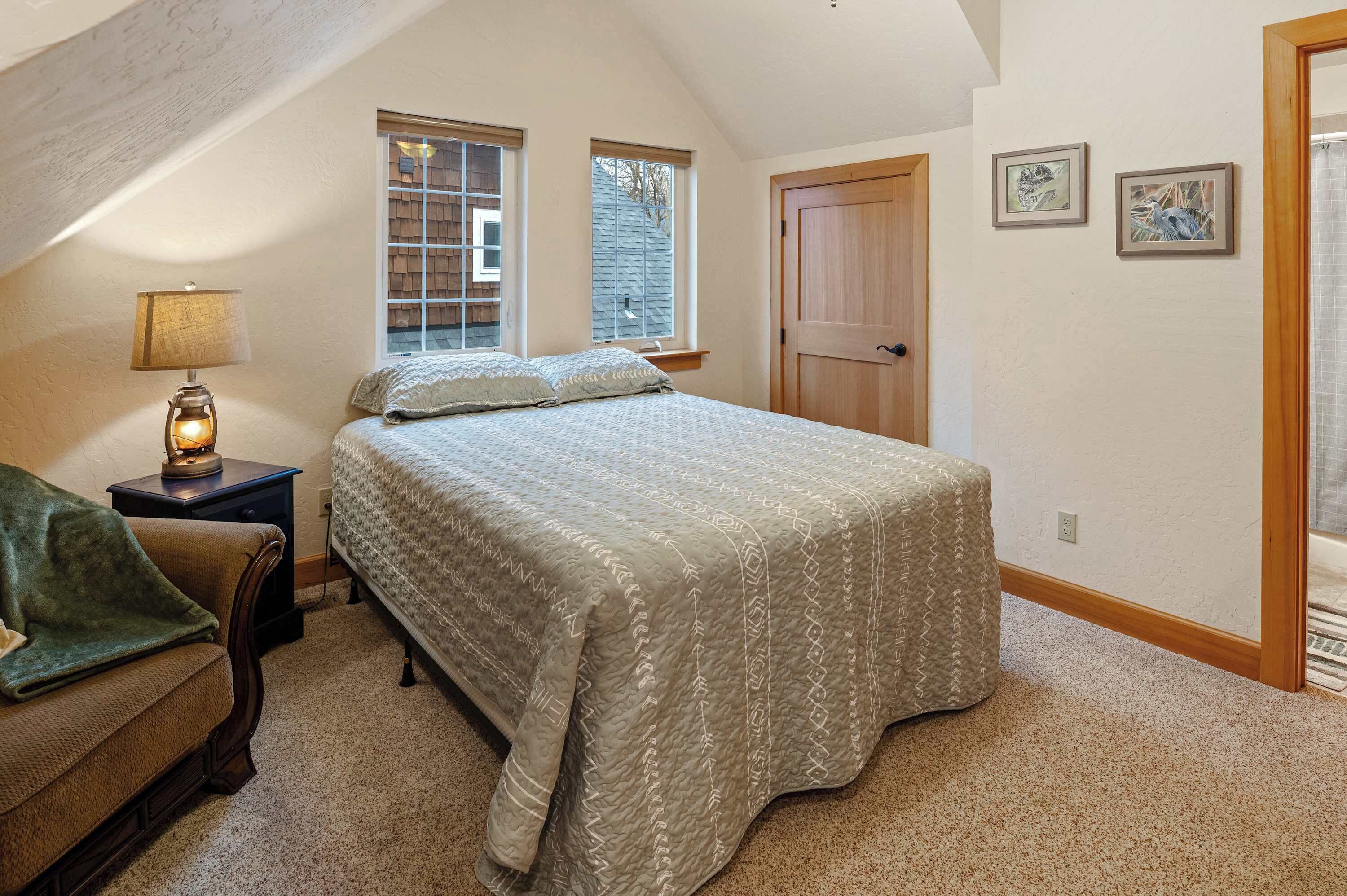 Une chambre à coucher avec un lit, une table de chevet avec une lampe, une chaise, deux fenêtres, une porte et deux tableaux encadrés accrochés au mur.