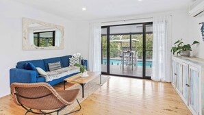 Ein modernes Wohnzimmer mit einem blauen Sofa, einem Korbstuhl, einem Holztisch und Blick auf einen Außenpool durch große Glaswände.