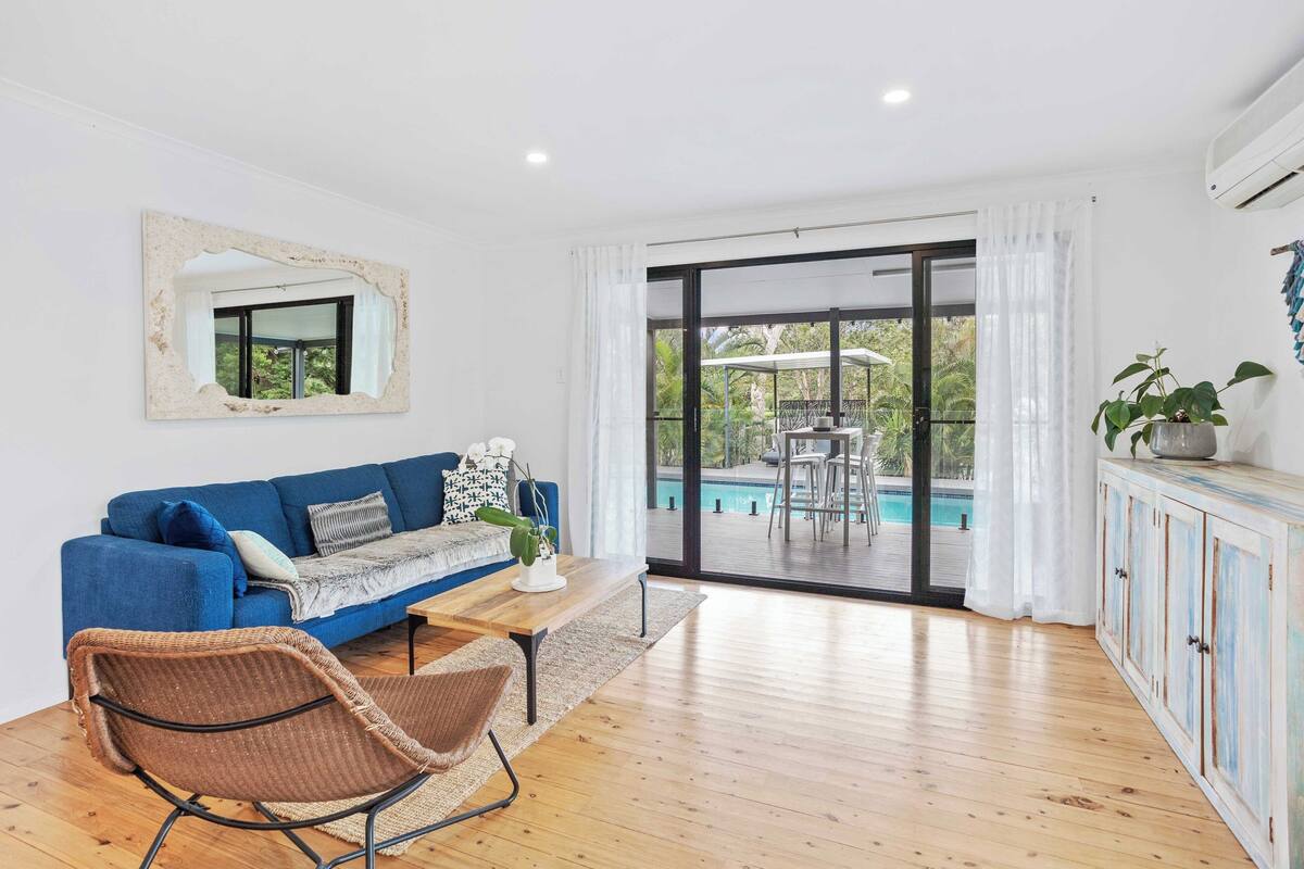 A modern living room with a blue sofa, a wicker chair, a wooden coffee table, and a view of an outdoor pool through large glass doors.
