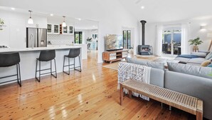 A modern kitchen with white cabinets, a central island with bar stools, a dining area with a sofa and bench, and a living room with a fireplace. - Escape to Alawara (Tallai)