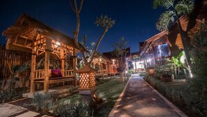A pathway leading to a wooden building with a thatched roof, surrounded by tropical plants and a well-lit garden area. - The Lebak Canggu (Canggu)