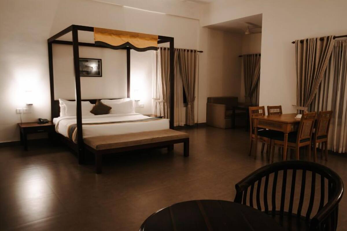A bedroom with a four-poster bed, a bedside table with a lamp, a wooden bench, a dining table with chairs, and curtains.