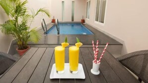 A poolside area with a table set for two, featuring two drinks and a glass of water, surrounded by potted plants and wicker chairs. - Abad Brookside (Vythiri)
