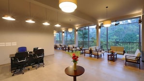A spacious room with a long table and chairs, large windows offering a view of the outdoors, and a ceiling with multiple hanging lights. - Abad Brookside (Vythiri)