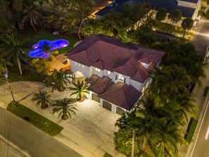 A two-story house with a swimming pool, surrounded by palm trees and a well-lit driveway.