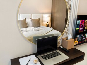 A hotel room with a bed, a laptop on a desk, a remote control, and a TV displaying movie options.