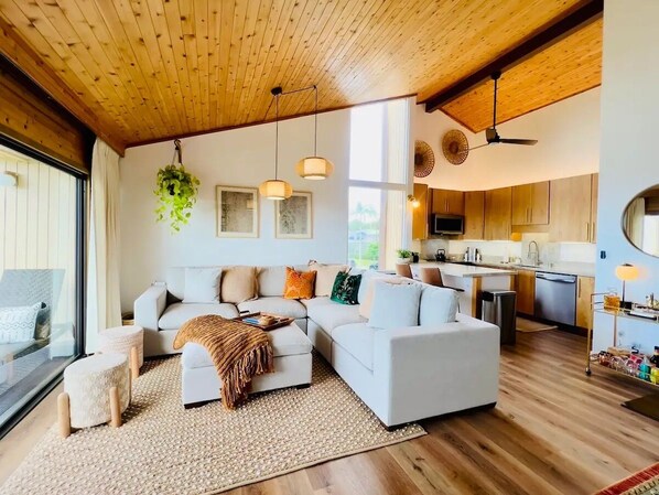 A modern living room with a large sectional sofa, a coffee table, and a dining area with wooden cabinets and a kitchen in the background.