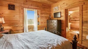 A wooden cabin bedroom with a bed, dresser, television, and a door leading to a bathroom. - Rustic Barn on 51 Acres (Cripple Creek)