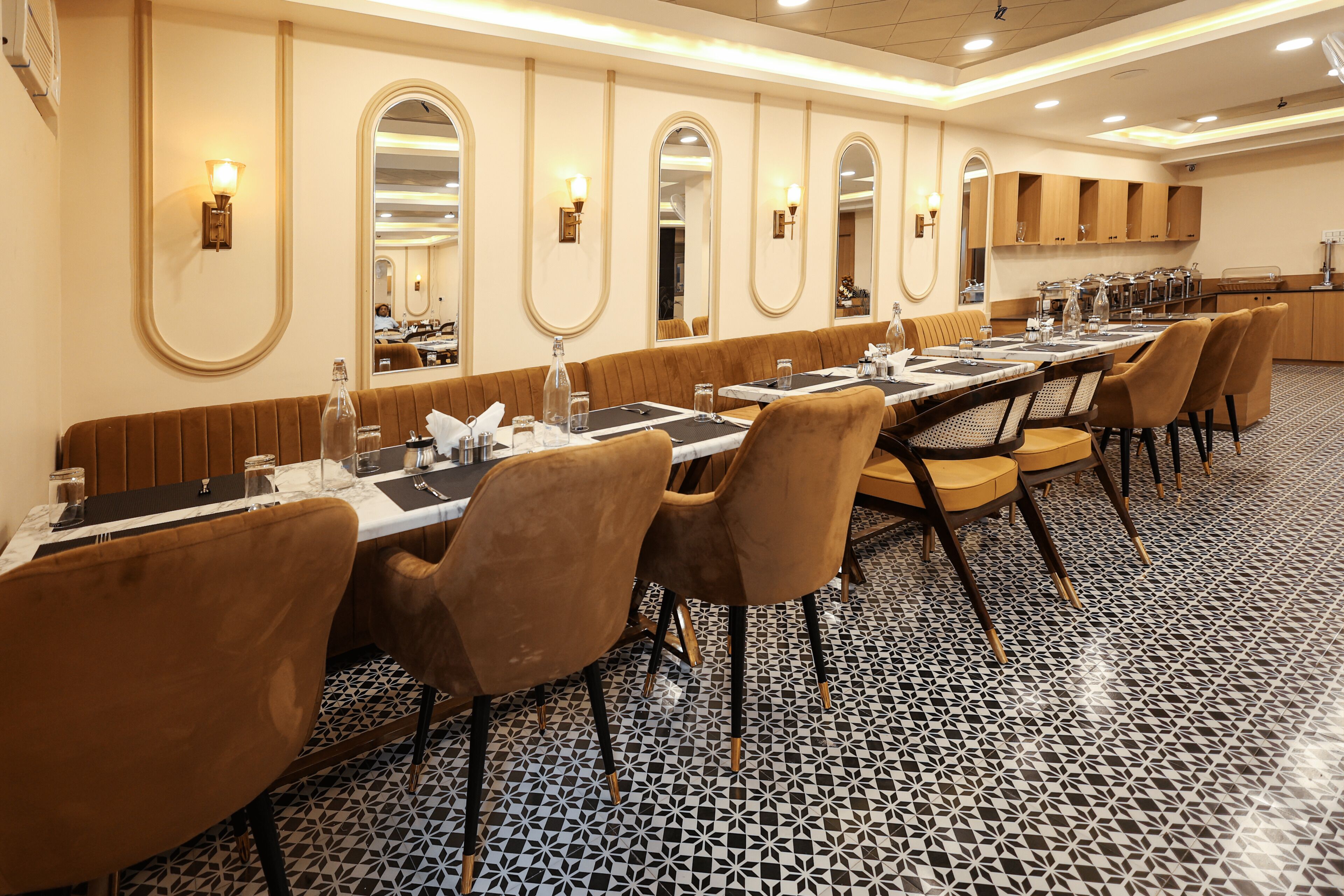 A long dining table with multiple chairs, a patterned floor, and a kitchen area in the background.