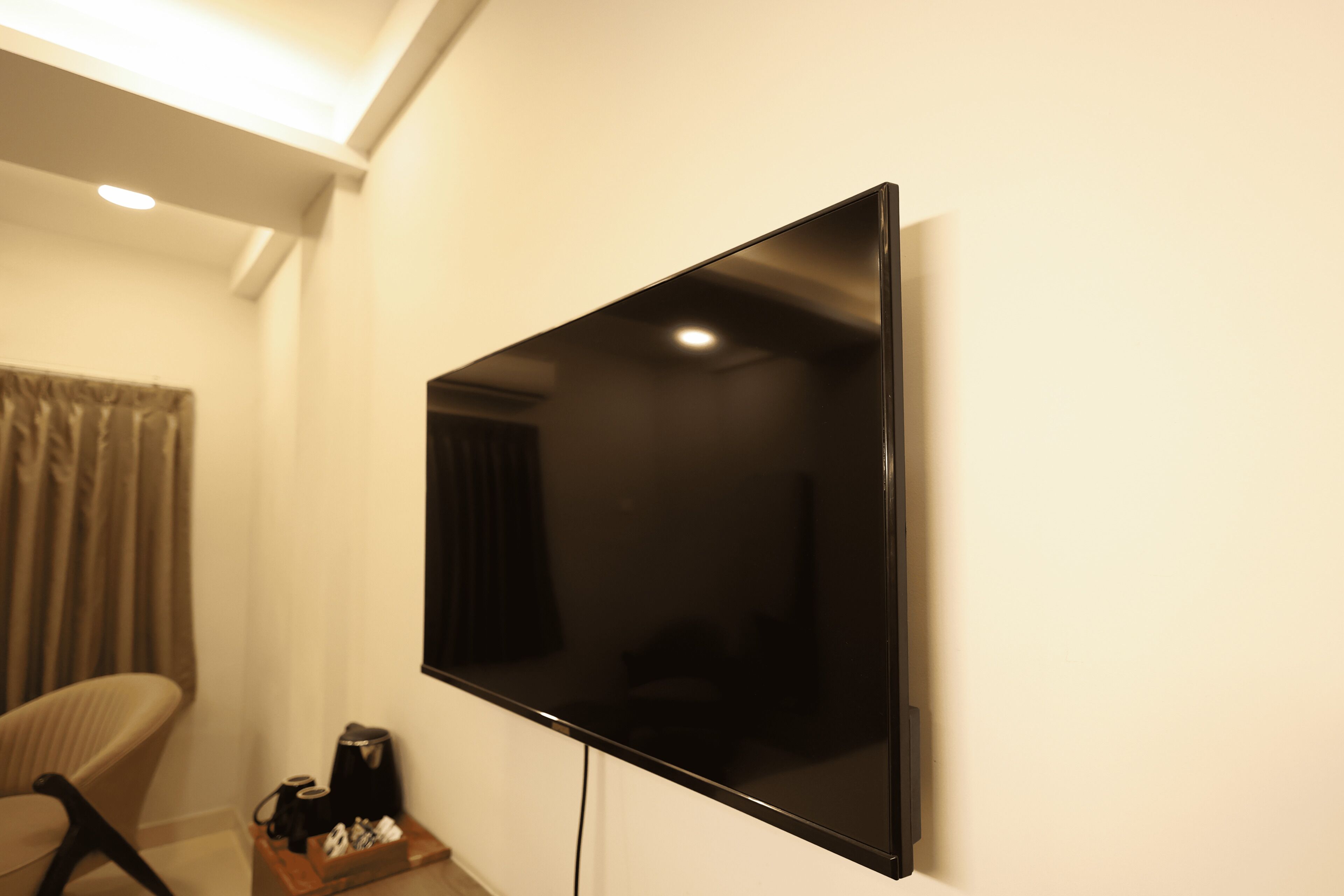 A flat-screen TV mounted on a wall in a room with a chair and a small table with a coffee maker.