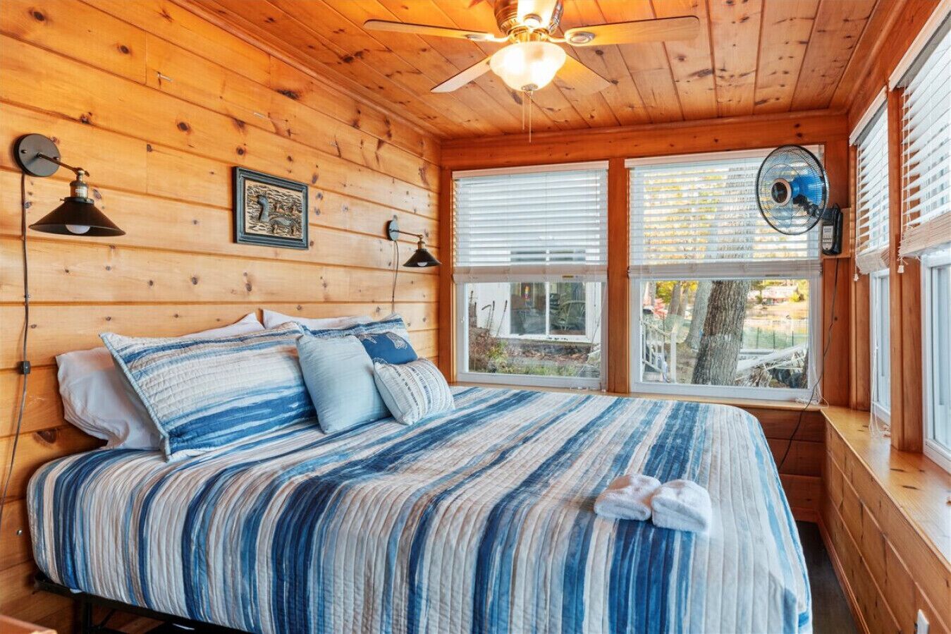 Dormitorio de cabaña de madera con una cama, dos lámparas de pared, un ventilador de techo y una ventana con persianas.