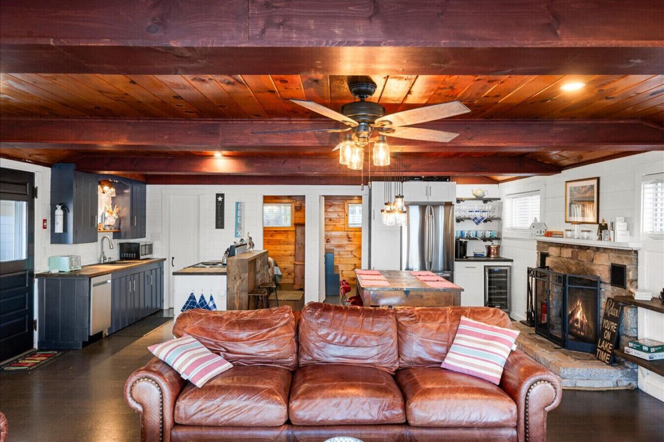 A spacious living area with a leather sofa, wooden ceiling beams, a ceiling fan, and a fireplace.