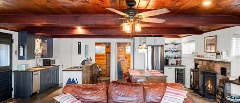 A spacious living area with a leather sofa, wooden ceiling beams, a ceiling fan, and a fireplace.