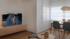 A modern living room with a flat-screen TV mounted on a wooden console, a dining area with a table and chairs, and a large window with sheer curtains. - Sunstay Cosy Flat I Cristo Malaga (Málaga)