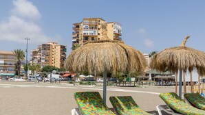 Une plage avec des chaises longues et des parasols en chaume, un espace de stationnement et, au loin, des immeubles à plusieurs étages.