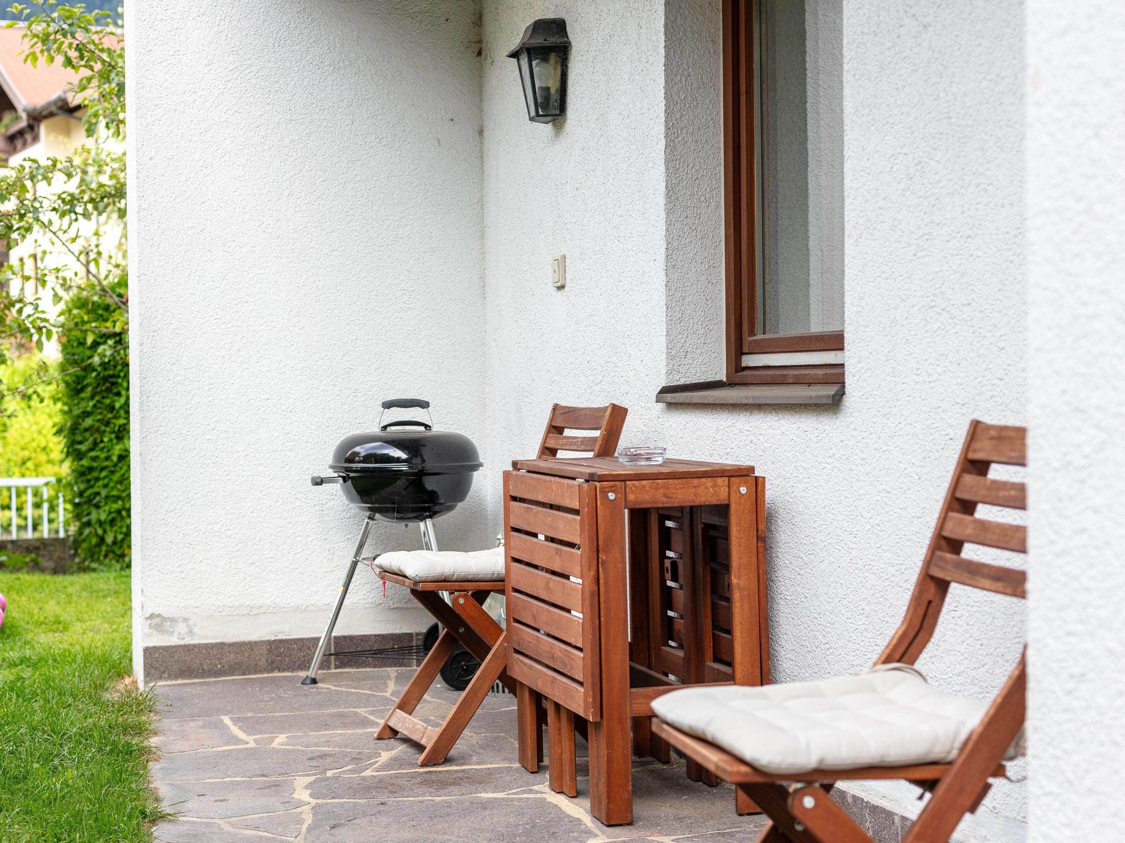 Un patio al aire libre con una mesa y sillas de madera, una parrilla y una ventana.