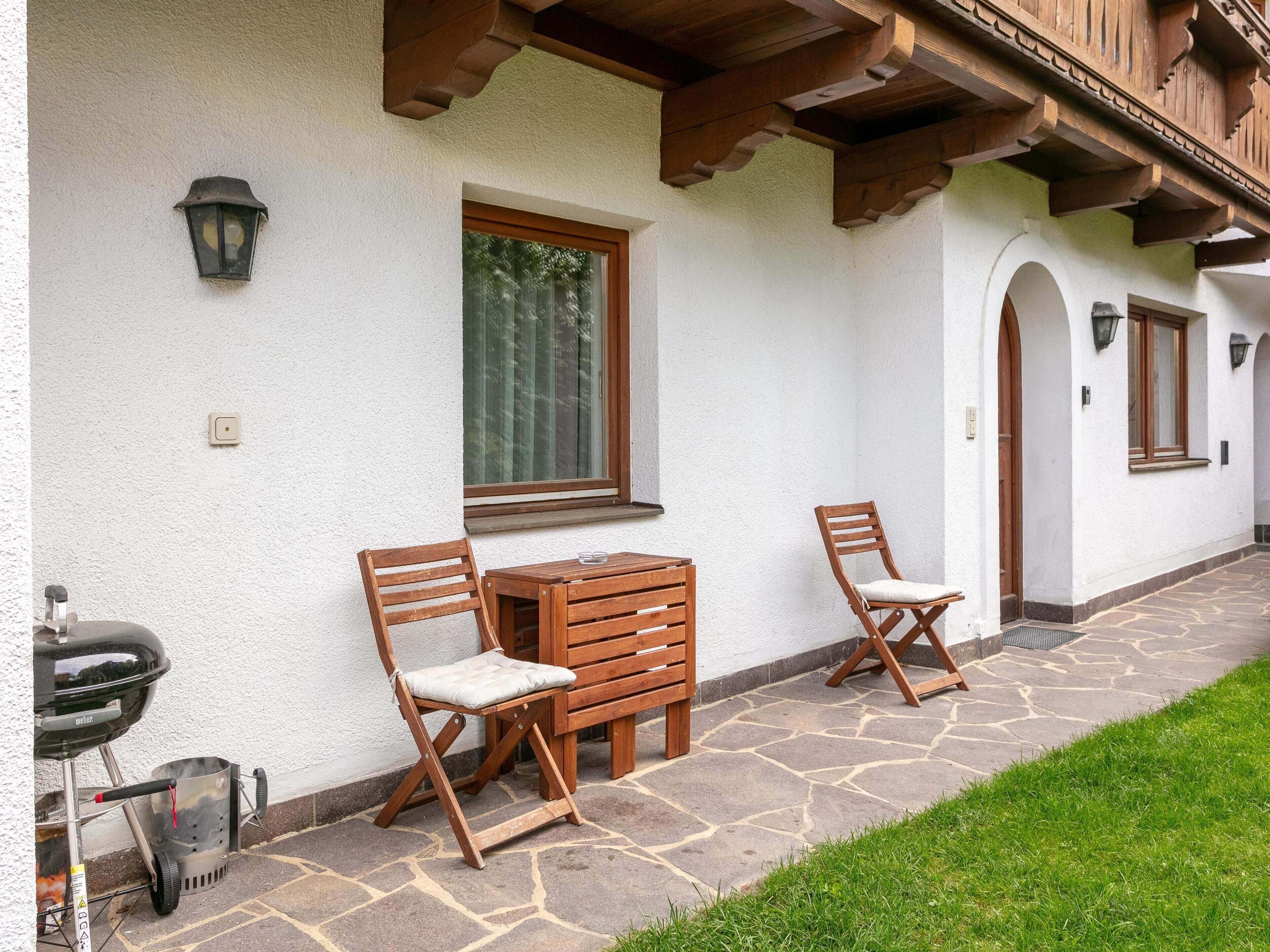 A patio with two wooden chairs and a table, a barbecue grill, and a stone pathway leading to a building with wooden beams and a white exterior.