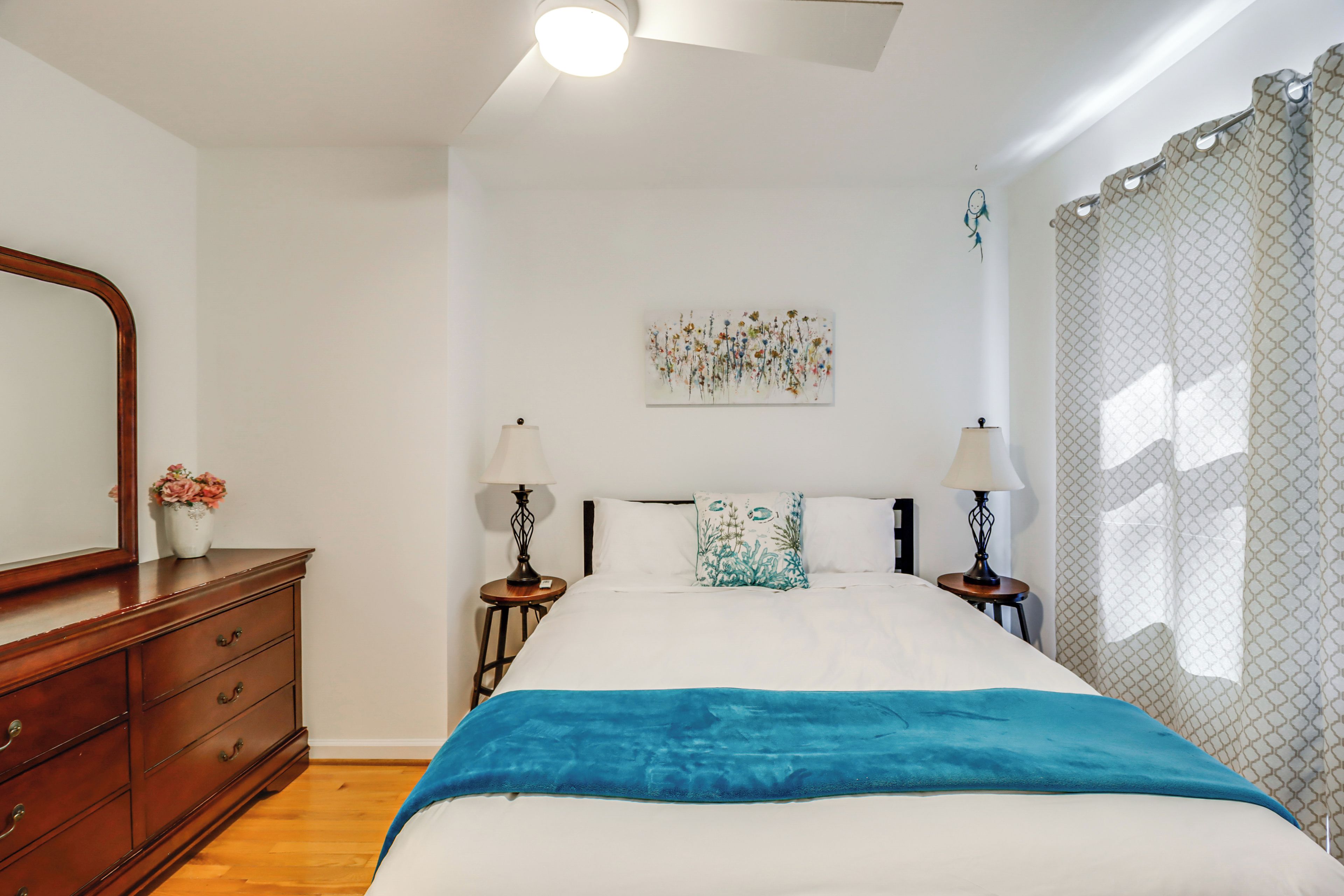 A bedroom with a bed, wooden dresser, two bedside tables with lamps, a wall art piece, and a window with curtains.