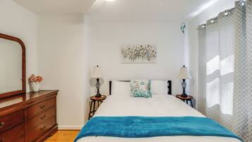 A bedroom with a bed, wooden dresser, two bedside tables with lamps, a wall art piece, and a window with curtains.