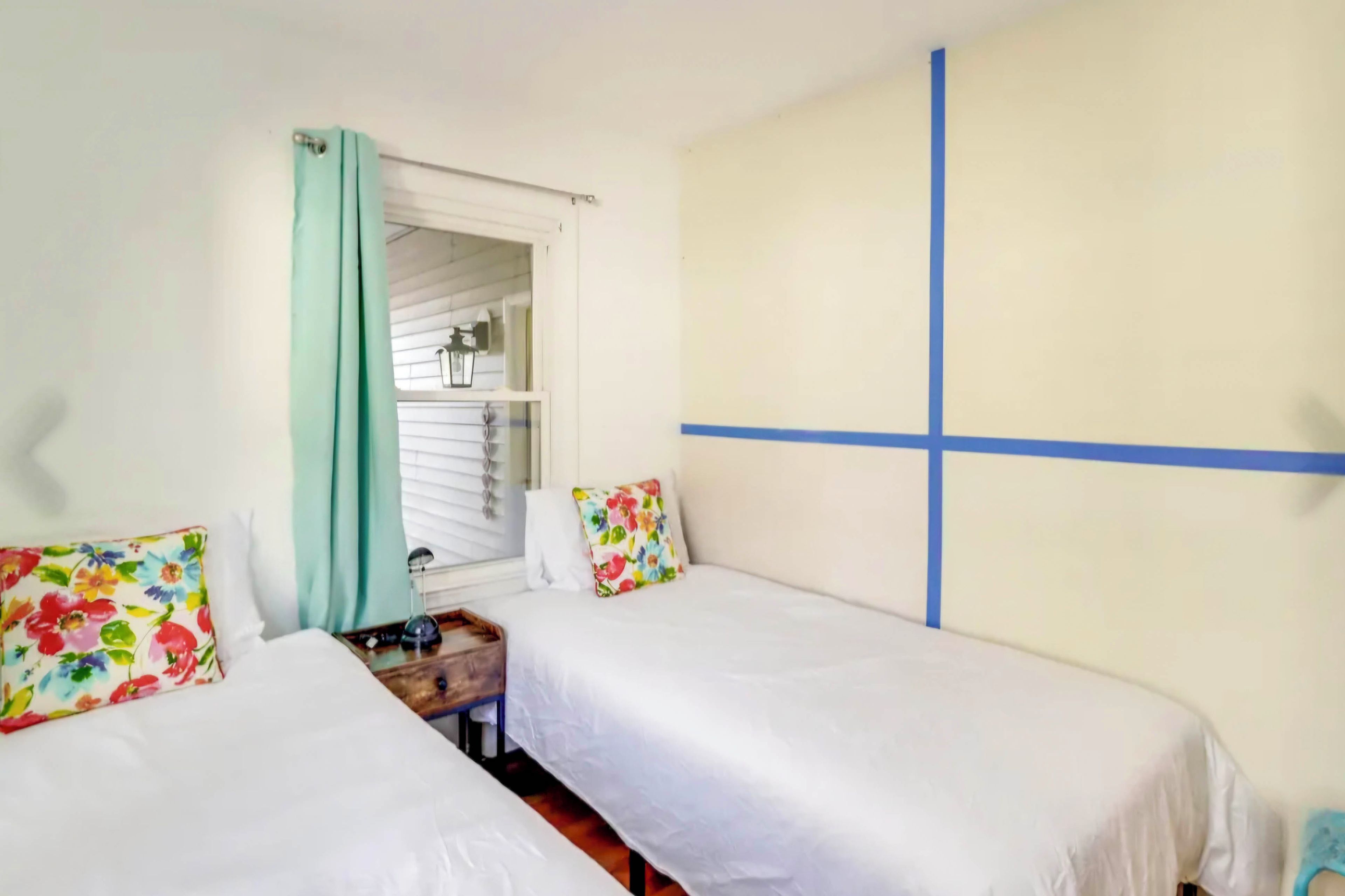 A room with two beds, a window with curtains, a small table, and a wall with a blue stripe.