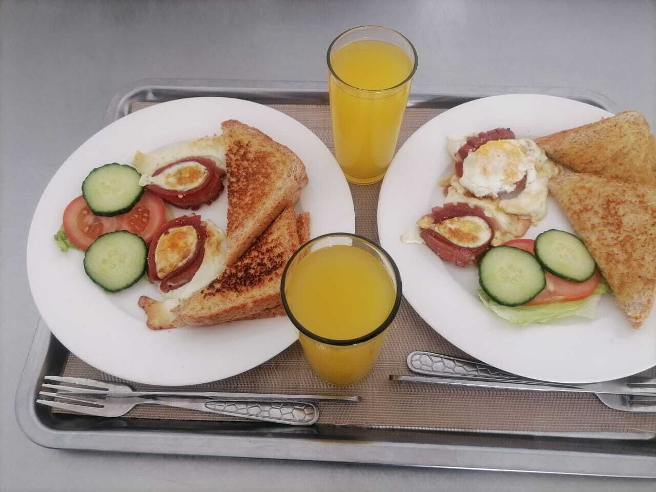 Uma bandeja com duas porções de café da manhã, cada uma com torradas, ovos, bacon, tomates e pepinos, e dois copos de suco de laranja.