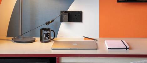 A modern desk with a laptop, a mug, and a notebook. There is a teddy bear on a shelf and a potted plant.