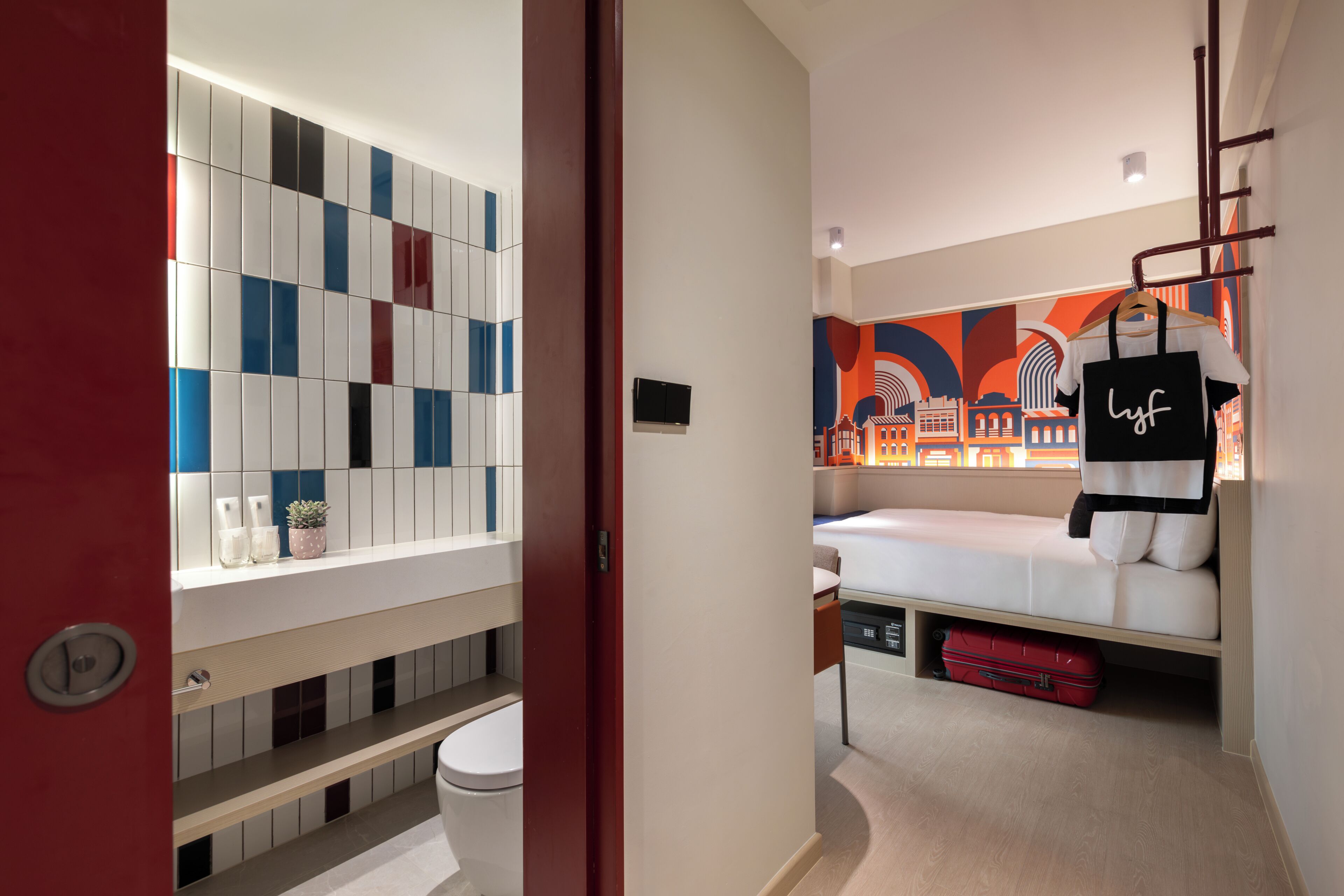 Moderni hotellihuone, jossa on sänky, punainen matkalaukku, seinä, jossa on kaupunkimaisemaa esittävä seinämaalaus, ja laatoitettu seinä, jossa on geometrinen kuvio.