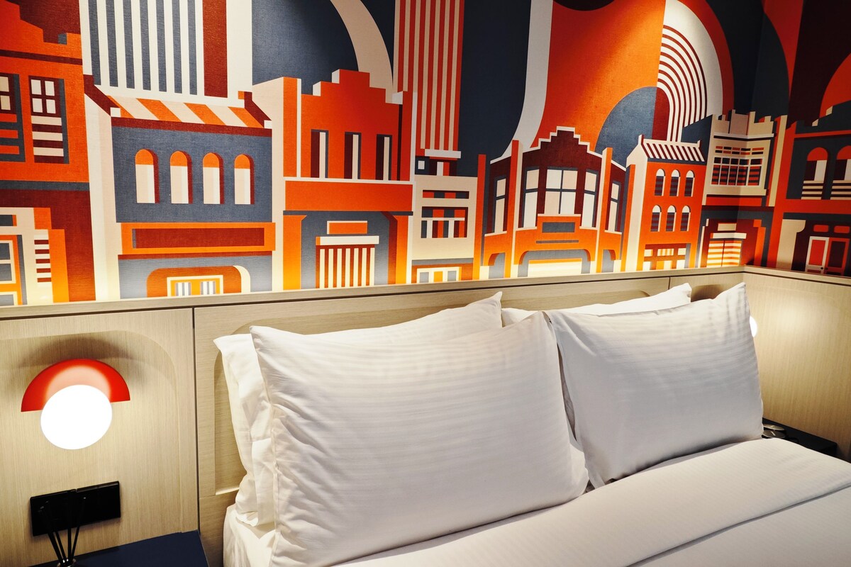 Quarto de hotel com cama, almofadas brancas, mesa de cabeceira, luminária na parede e um mural com vista urbana.