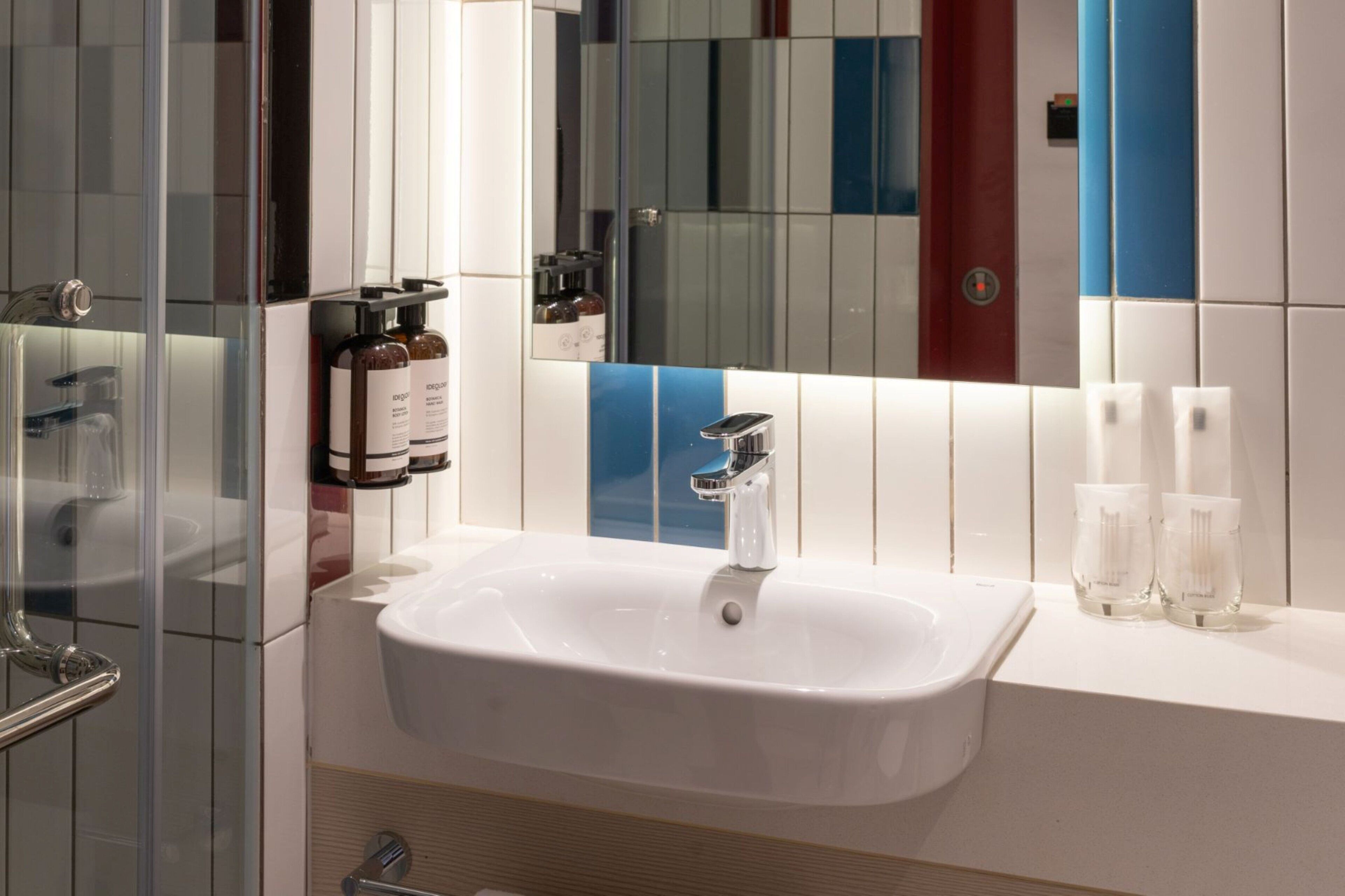 Un bagno moderno con lavandino bianco, box doccia in vetro e uno specchio che riflette una parete colorata.