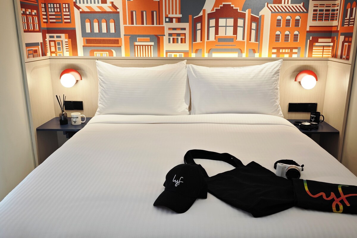 A hotel room with a large bed, two bedside tables, a nightstand, a lamp, a mug, a cap, and a bag.