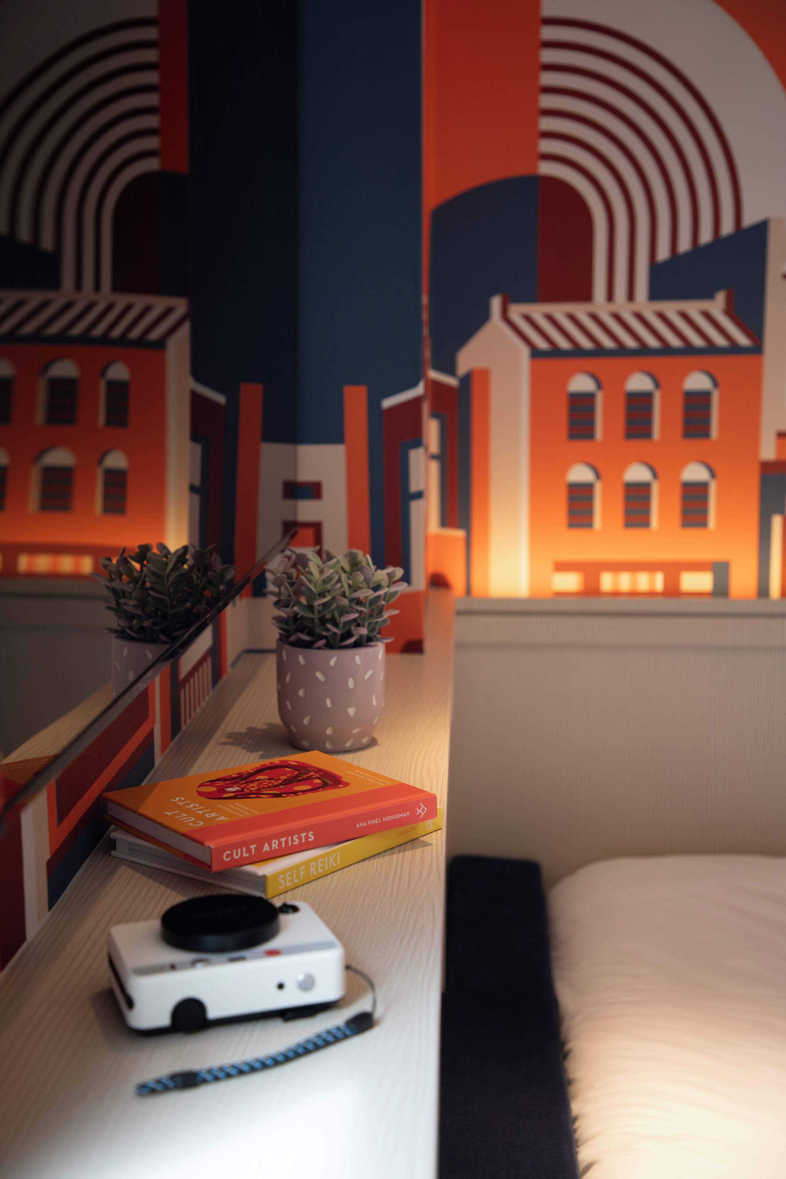 A neatly arranged desk with a camera, books, and potted plants against a backdrop of a stylized cityscape mural.