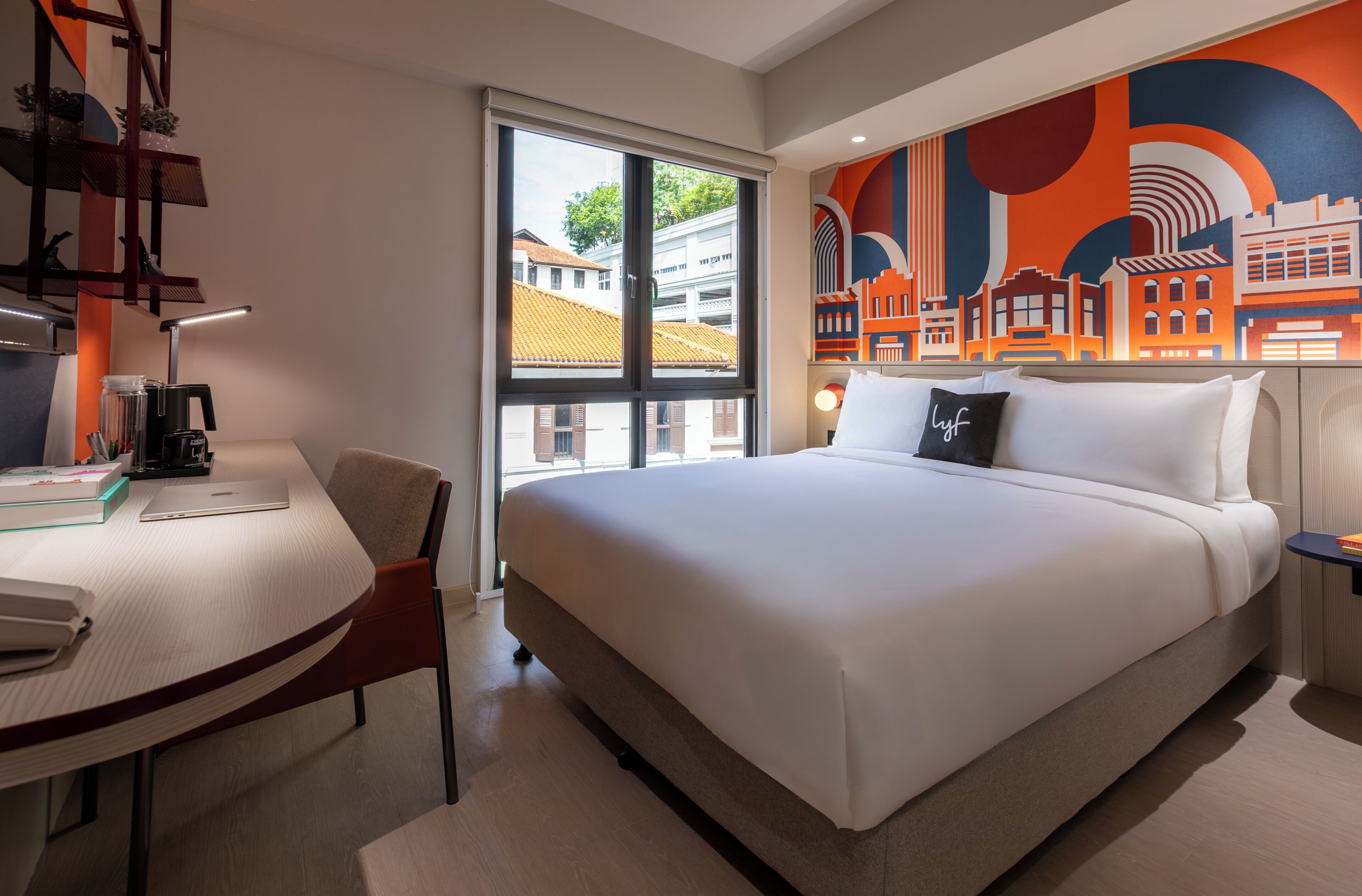 Quarto de hotel com cama, escrivaninha, cadeira, janela e um mural com vista para a cidade.
