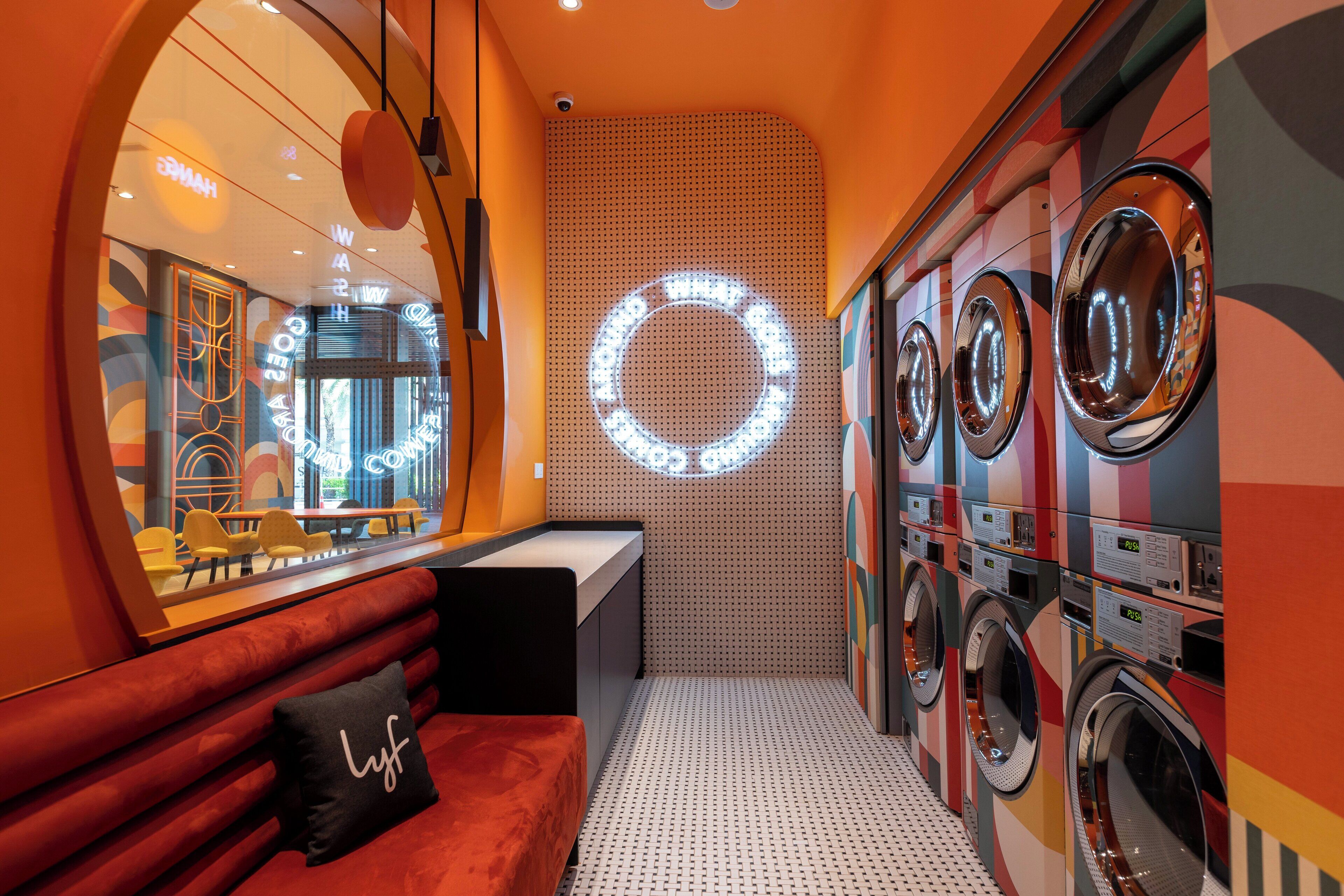 Une buanderie équipée de plusieurs machines à laver, un coin salon avec un banc rouge et un luminaire circulaire fixé au mur.