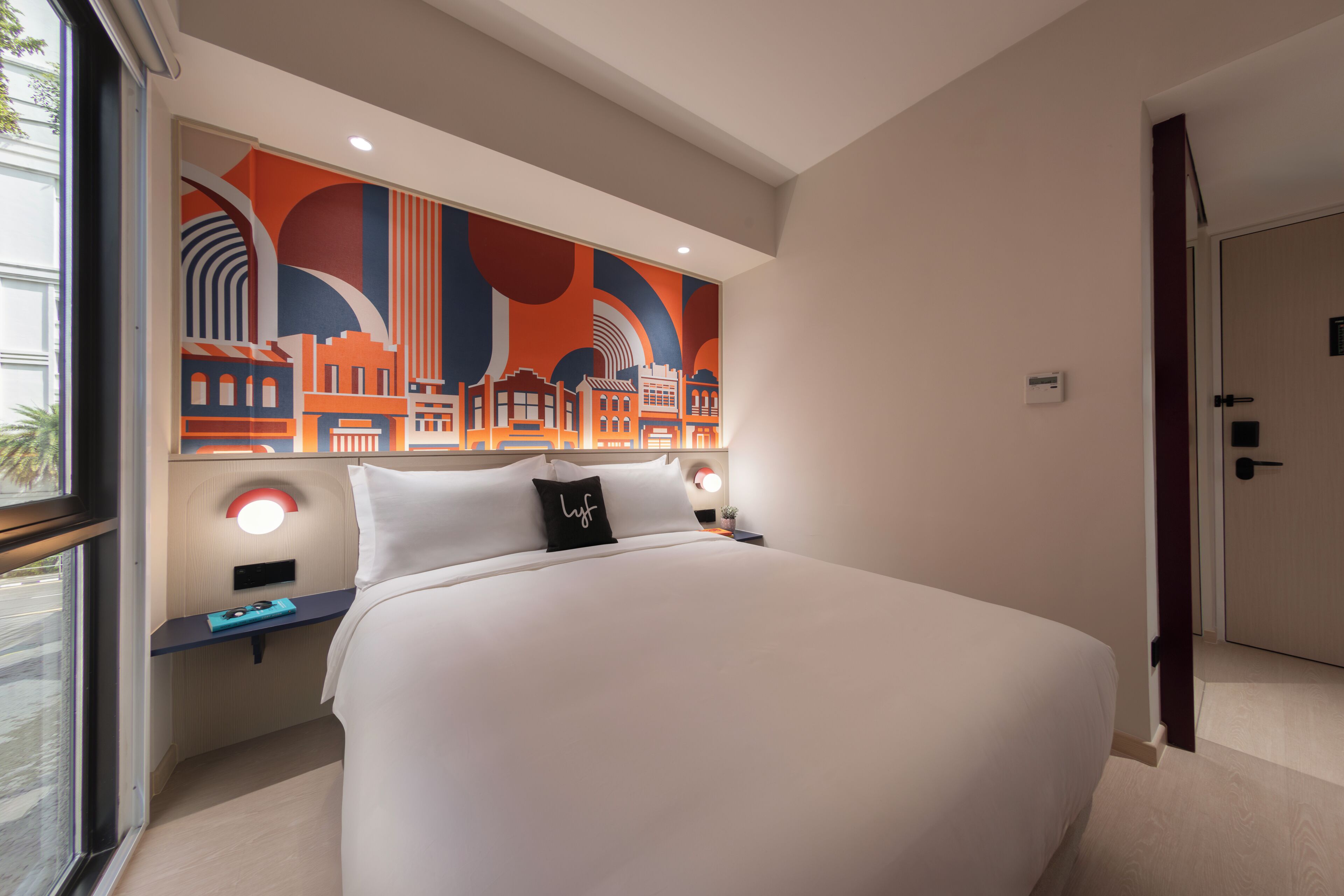 A hotel room with a large bed, a cityscape artwork on the wall, a bedside table with a lamp, and a door to the right.