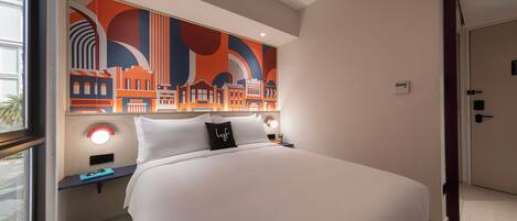 A hotel room with a large bed, a cityscape artwork on the wall, a bedside table with a lamp, and a door to the right.
