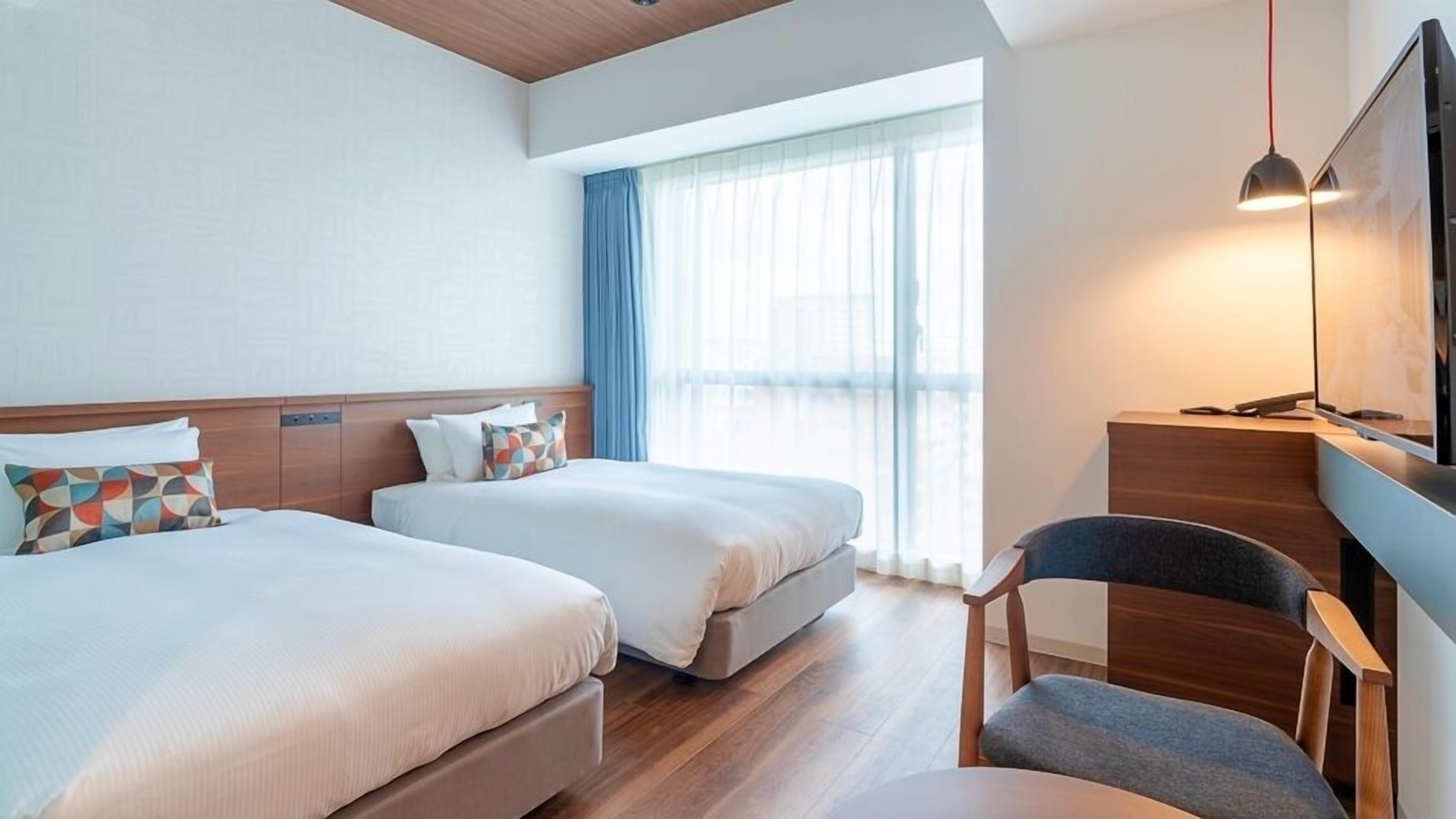 Een hotelkamer met twee bedden, een bureau, een stoel, een televisie en een groot raam met gordijnen.