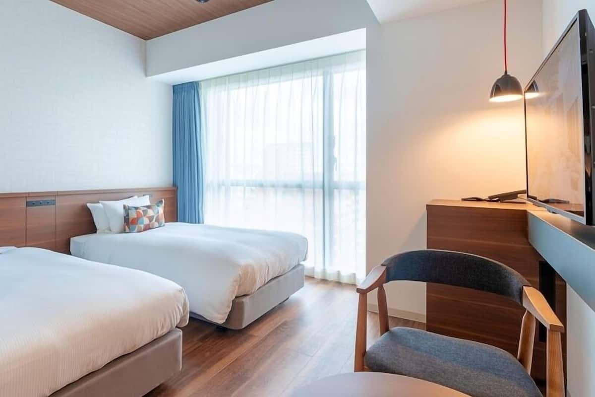 Quarto de hotel com duas camas, uma escrivaninha, uma cadeira, uma televisão e uma janela grande com cortinas.