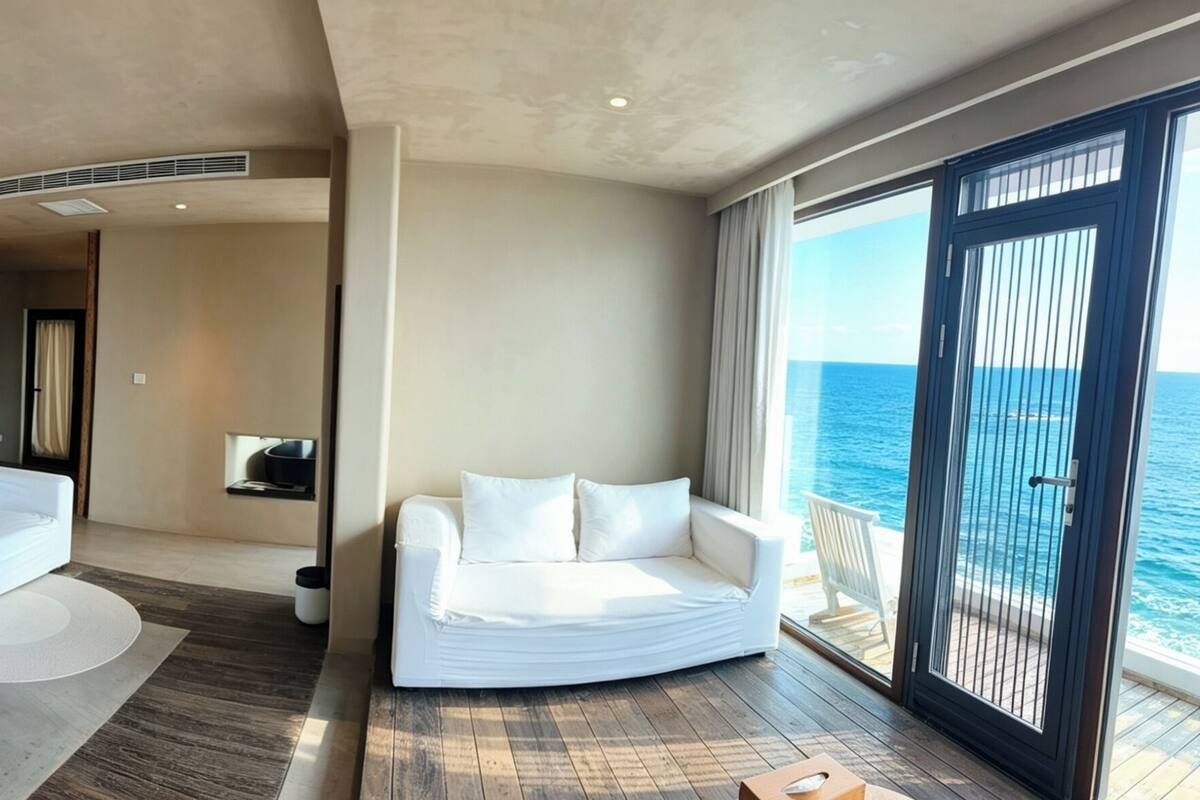 A hotel room with a bed, two white sofas, a wooden table with tea set, and a large window with a view of the sea.
