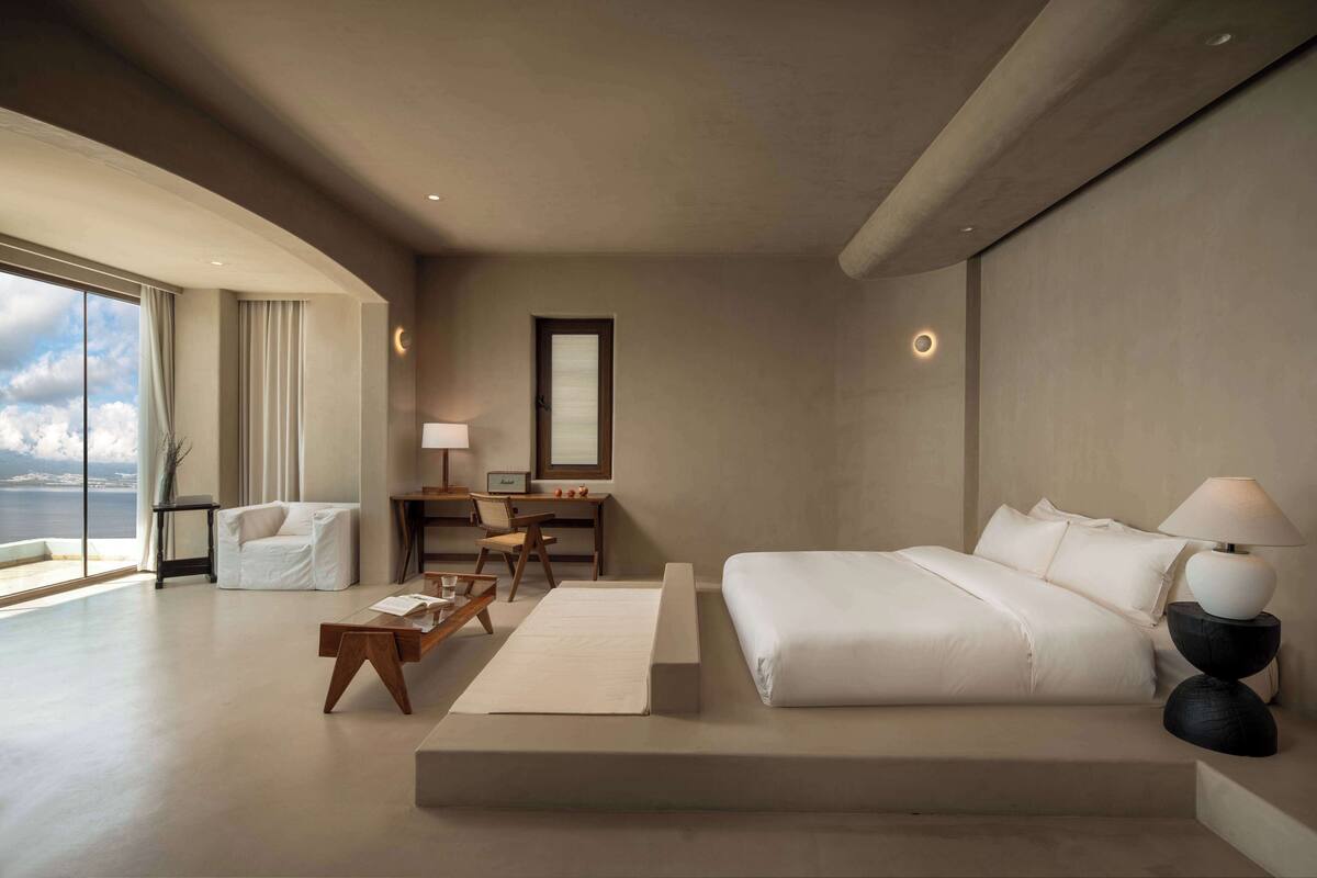 A modern hotel room with a large bed, a desk with a chair, a sofa, and a view of the sea.
