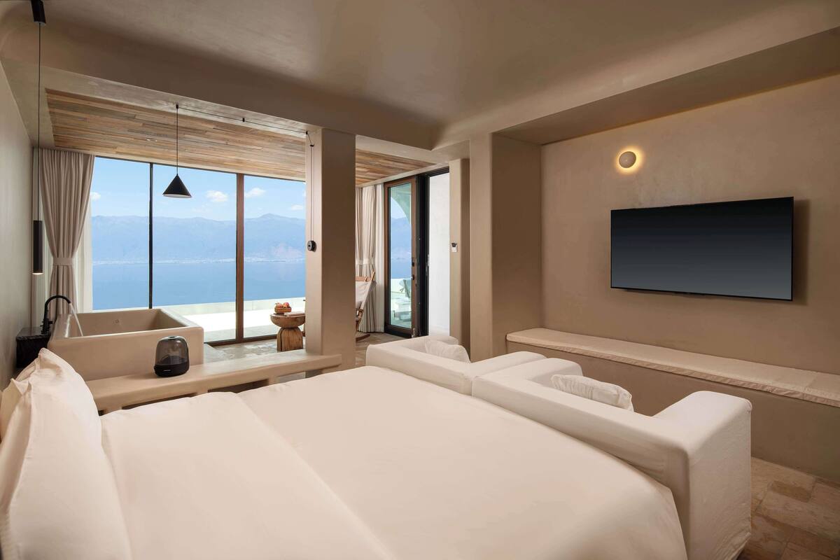 A modern hotel room with a large bed, a flat-screen TV mounted on the wall, a large window with a view of the sea, and a small table with a chair.