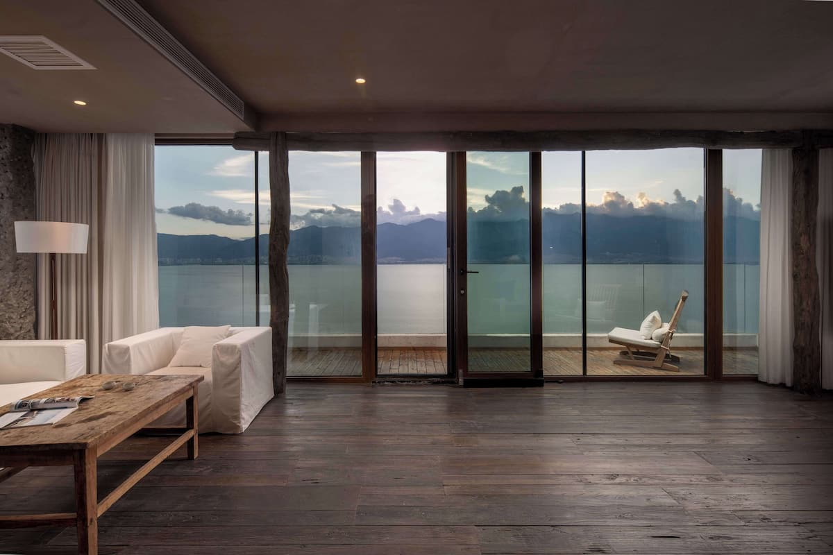 A spacious room with a large sliding glass door leading to a balcony with a view of a lake and mountains. There is a white sofa and a wooden table with a lamp.