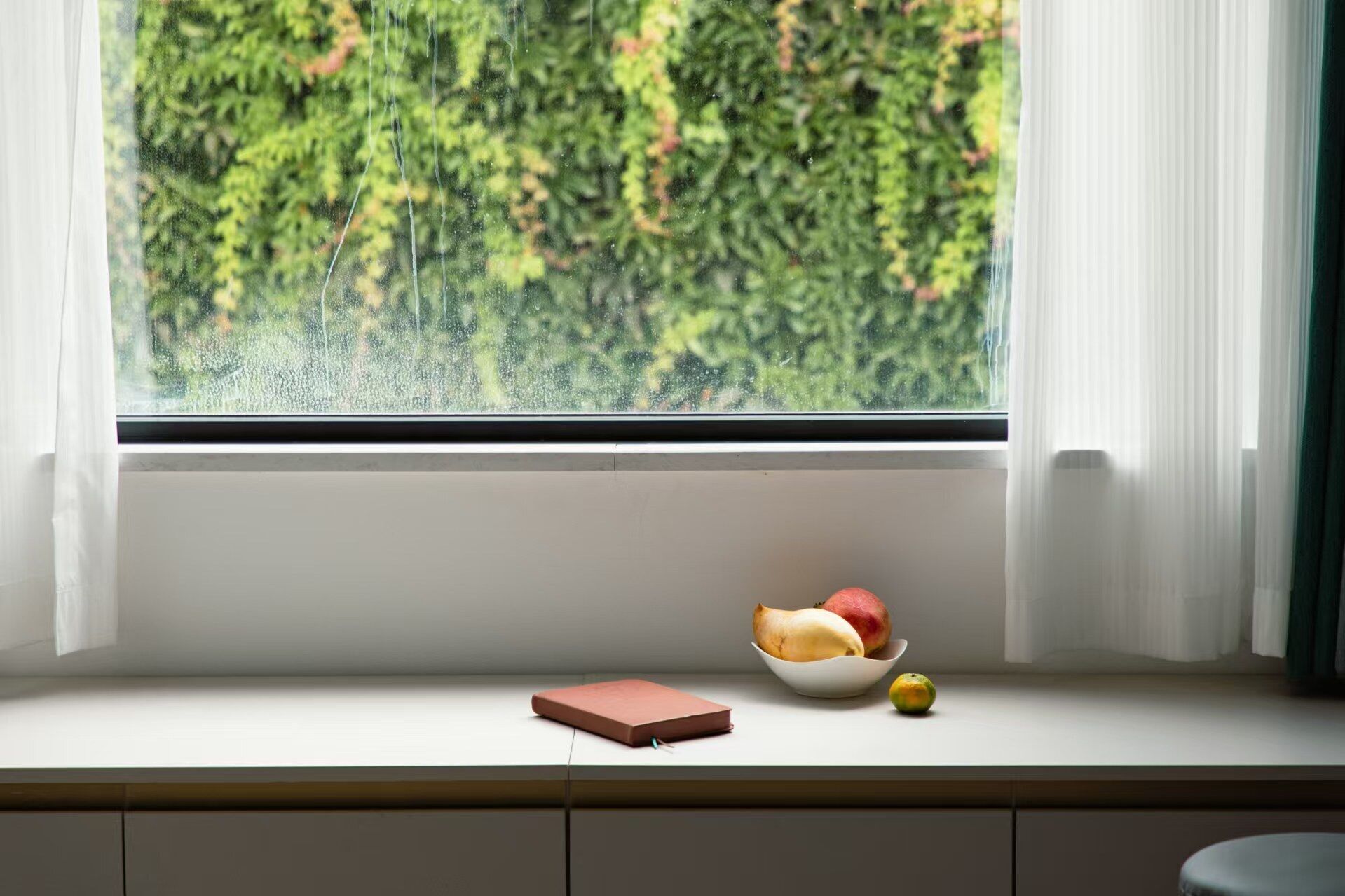 Una ventana con vistas a vegetación, un bol de fruta y una libreta sobre un repisa.